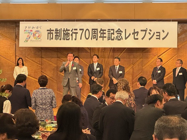 「市制施行70周年記念レセプション」と書かれた横断幕を掲げた舞台に複数の関係者が並んで立ち、前に出た高橋市長が、多くの人で賑わう会場に向かってマイクで話をしている写真