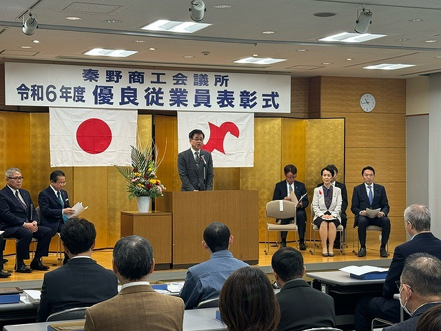「秦野商工会議所 令和6年度 優良従業員表彰式」の横断幕が掲げられ金屏風が建てられた舞台の左右に配置した椅子に複数の関係者が座り、中央の演台に立った高橋市長が、前の席に座った参加者たちに向かって話をしている写真