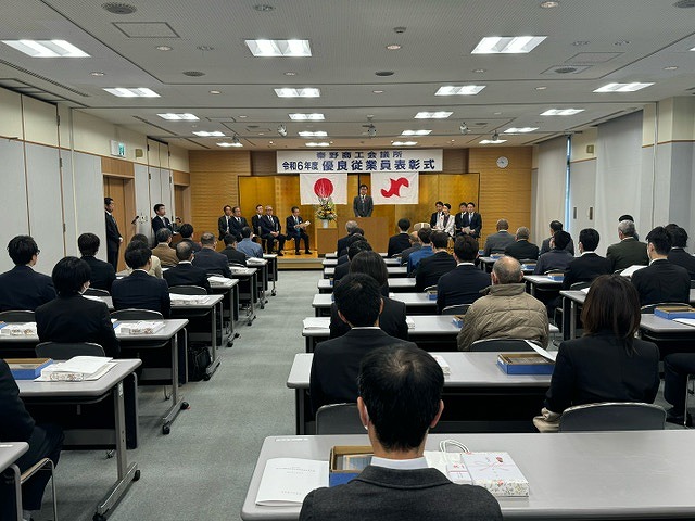 部屋の奥には金屏風を立てた舞台が設置され、たくさん並んだ長机の席には大勢の参加者たちが座って、登壇者の話を聞いている表彰式の様子を後方から撮影した写真