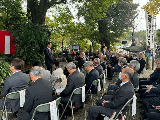 緑の木々に囲まれ、大きな石灯籠がある会場に並べられた椅子に座った大勢の参列者が、前方で話をしている高橋市長の方を見ている写真