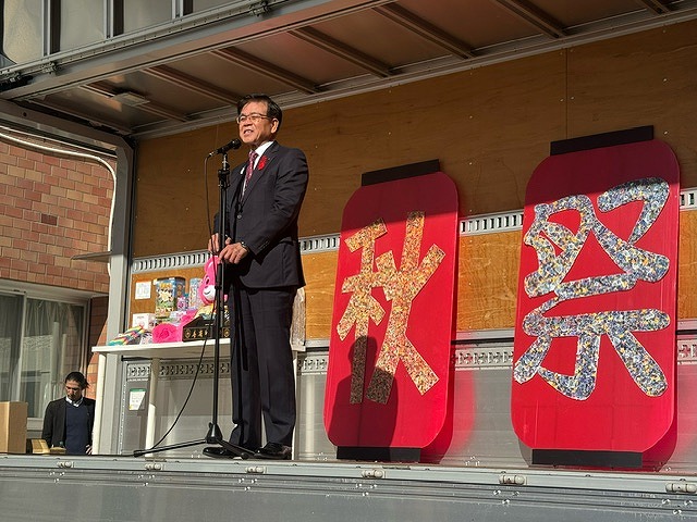 それぞれ「秋」「祭」と一文字ずつ文字が貼られた、赤い提灯の形をした大きな看板が2つ並び、その前に立った高橋社長が、スタンドマイクに向かって話をしている写真