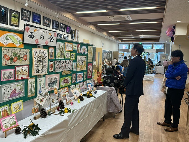 掲示板にたくさんの絵などが飾られ、その下に並んだテーブルに秋の植物や果実、小さいサイズの絵の作品などが並んだ展示スペースの前に立ち、作品を眺めている高橋市長と関係者の男性の写真