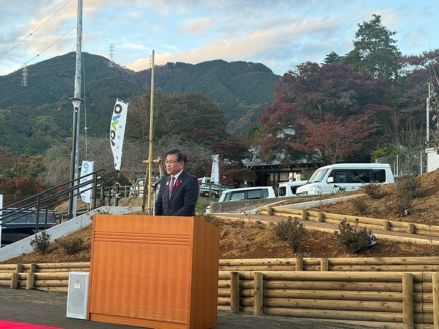 歩道が設けられたなだらかな斜面の上にある駐車場と奥の山を背景に、屋外に置かれた演台に立った高橋市長がスピーチをしている様子の写真