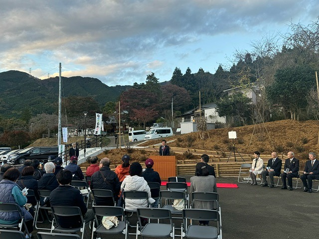 駐車場と山を背景に、演台に立った高橋市長がスピーチをしており、右側の椅子に4人の関係者が座り、手前に並んだ椅子には複数の人々が座っている様子の写真