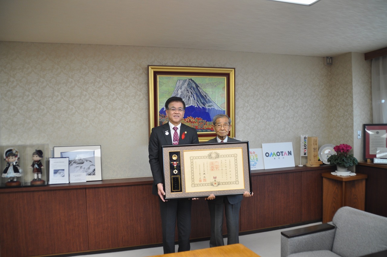 山の絵画が掛けられた壁を背に、高橋市長と佐野友保さんが並んで立ち、勲章と賞状が入った大きな額を一緒に持って写した記念写真