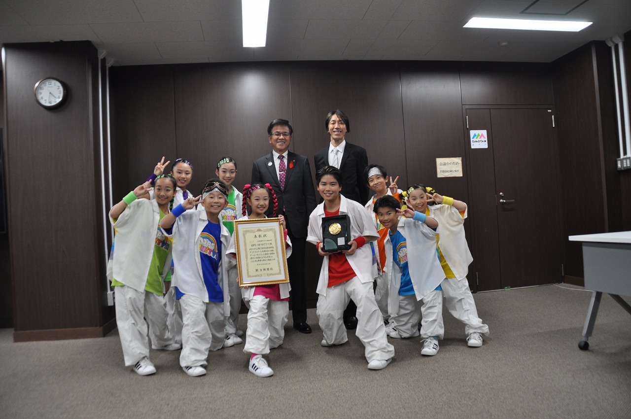 笑顔で並んで立った高橋市長と関係者の男性の前に、賞状と盾を持った2人を中央にして、白を基調としたおそろいの衣装を着た9人の子どもたちが並び、ピースサインをして写した記念写真