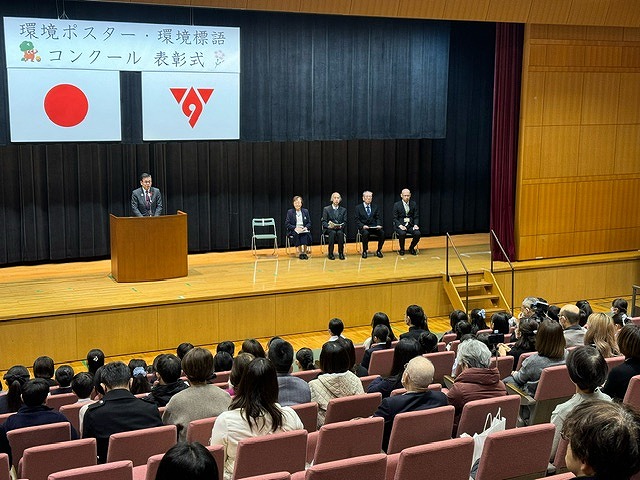 「環境ポスター・環境標語コンクール表彰式」と書かれた横断幕と日本国旗、秦野市市章が掲げられた舞台の演台に立った高橋市長が、大勢の人々が着席した観客席に向かって話をしており、その横に並んだ椅子には4人の男女が着席している様子の写真