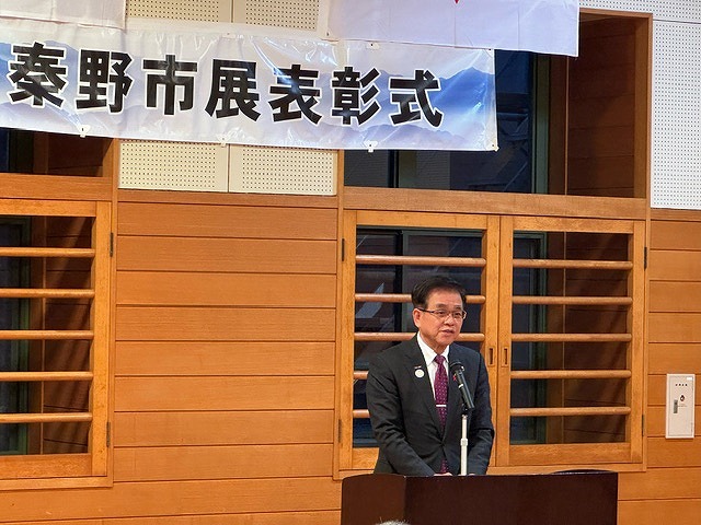 「秦野市展表彰式」と書かれた横断幕が掲げられた壁の前で、演台に立ちマイクに向かって話をしている高橋市長の写真