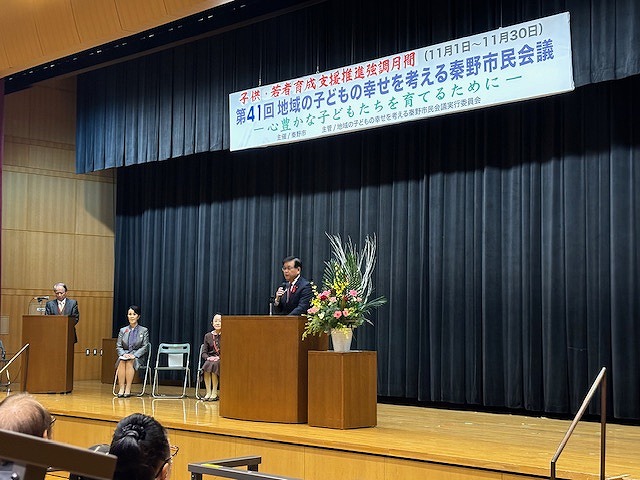 「第41回地域の子どもの幸せを考える秦野市民会議」の横断幕が掲げられた舞台の中央に置かれた、豪華な花が飾られた演台に立った高橋市長が、マイクを持って話をしており、後方の椅子に2人の女性が座っている様子の写真