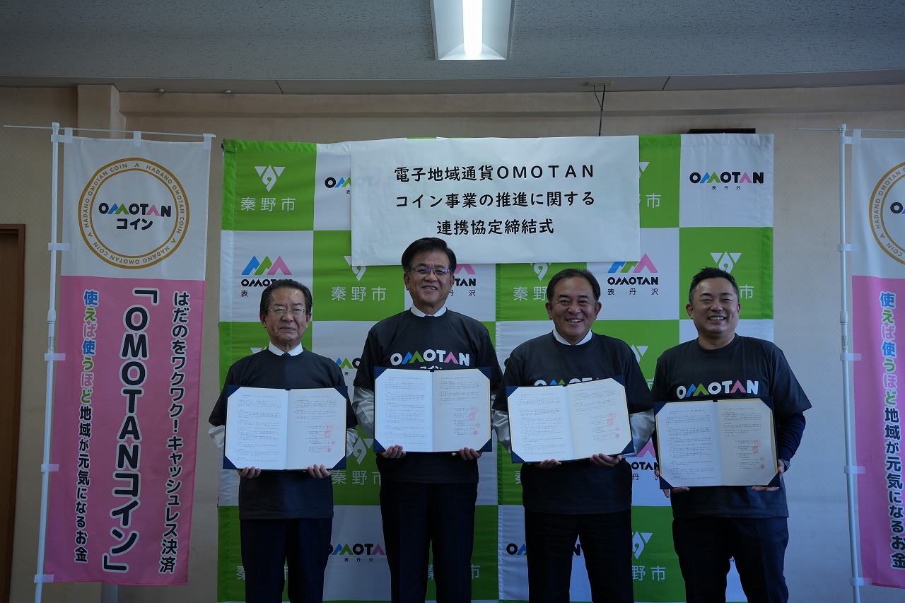 「OMOTANコイン」と書かれたピンク色ののぼりと、「電子地域通貨OMOTANコイン事業の推進に関する連携協定締結式」の横断幕が貼られたバックボードの前に、高橋市長を含む4人の男性が、おそろいのTシャツを着て、協定書を胸の前に持って並んで立っている記念写真