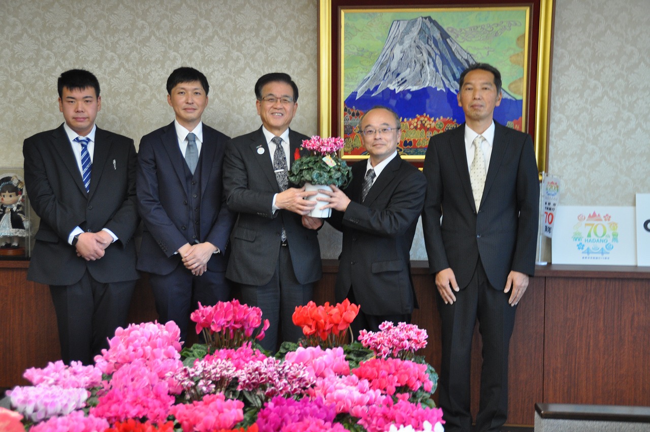 山の絵画が掛けられた壁を背に、ピンク色のシクラメンの鉢を持った高橋市長と男性、他、関係者の男性3人が並んで立ち、手前に置いたたくさんのシクラメンの花と一緒に写した記念写真