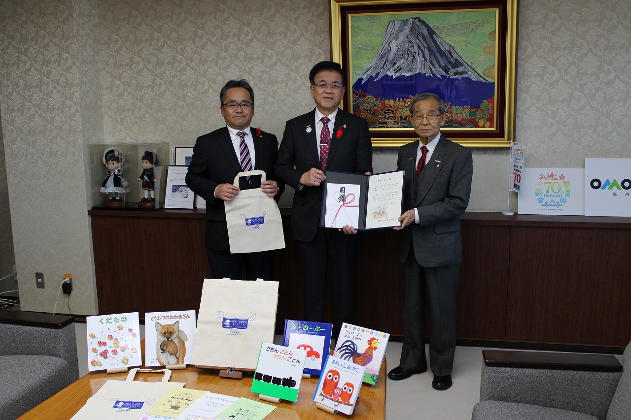 山の絵画が掛けられた壁を背に、目録やコットンバックを持った高橋市長と佐野代表取締役会長、関係者の男性が並んで立ち、手前のテーブル並べた複数の絵本やコットンバックと一緒に写した記念写真