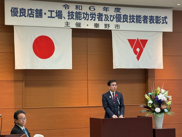 「令和6年度 優良店舗・工場・技能功労者及び優良技能者表彰式」の横断幕と日本国旗、市章が掲げられた壁の前で、演台に立った市長が話をしている写真