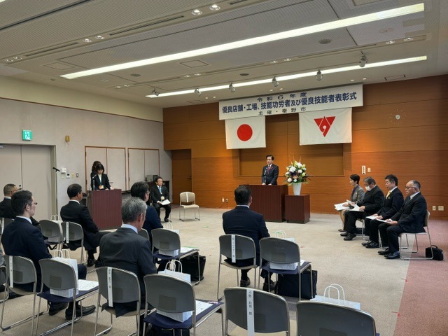「令和6年度 優良店舗・工場・技能功労者及び優良技能者表彰式」の横断幕と日本国旗、市章が掲げられた壁の前で、演台に立った市長が話をしており、会場に並んだ椅子には複数の出席者たちが座っている様子の写真