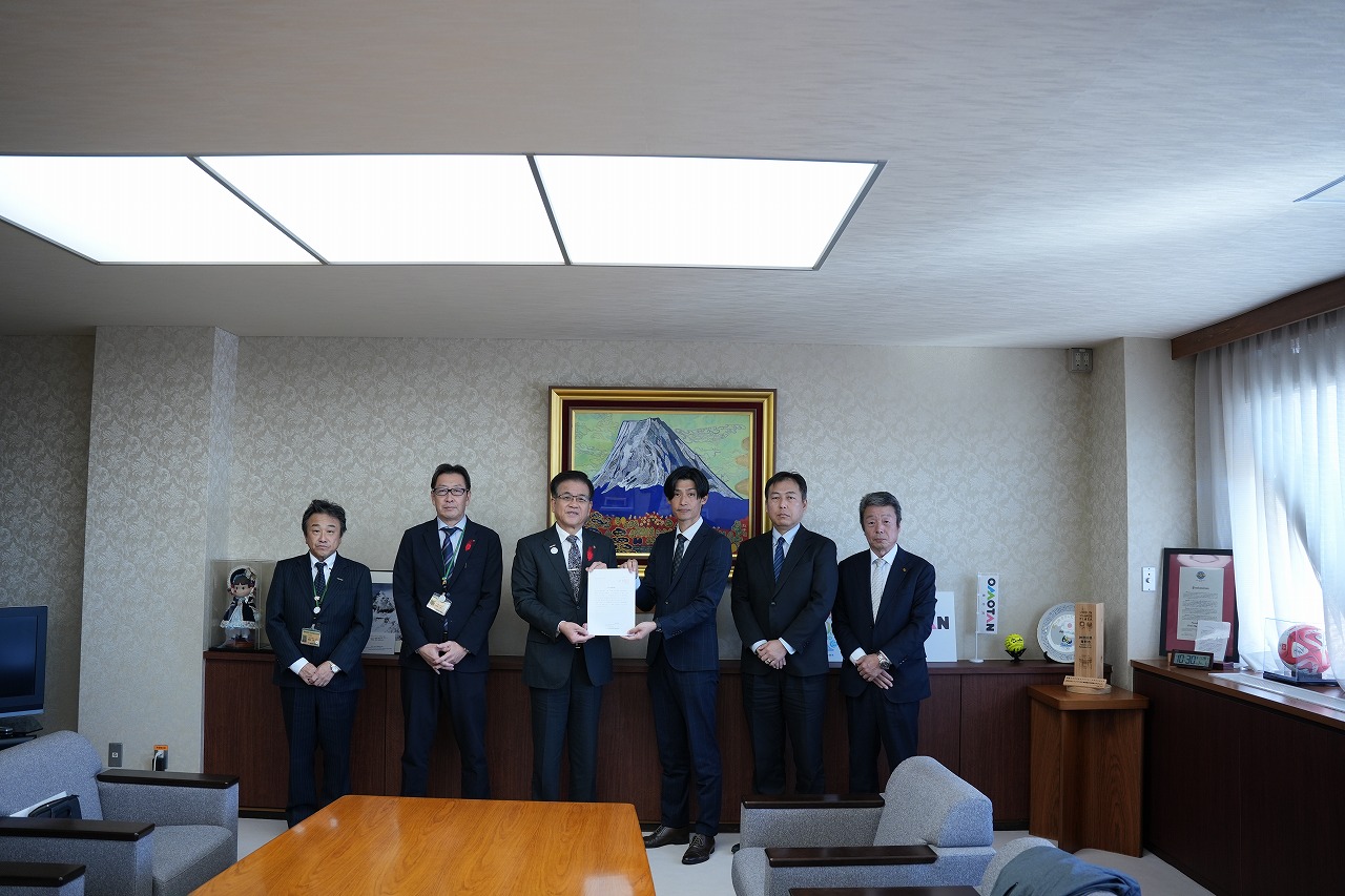 山の絵画が掛けられた壁を背に、要求書を持った高橋市長と男性を中心に、4人の関係者の男性が並んで立っている記念写真