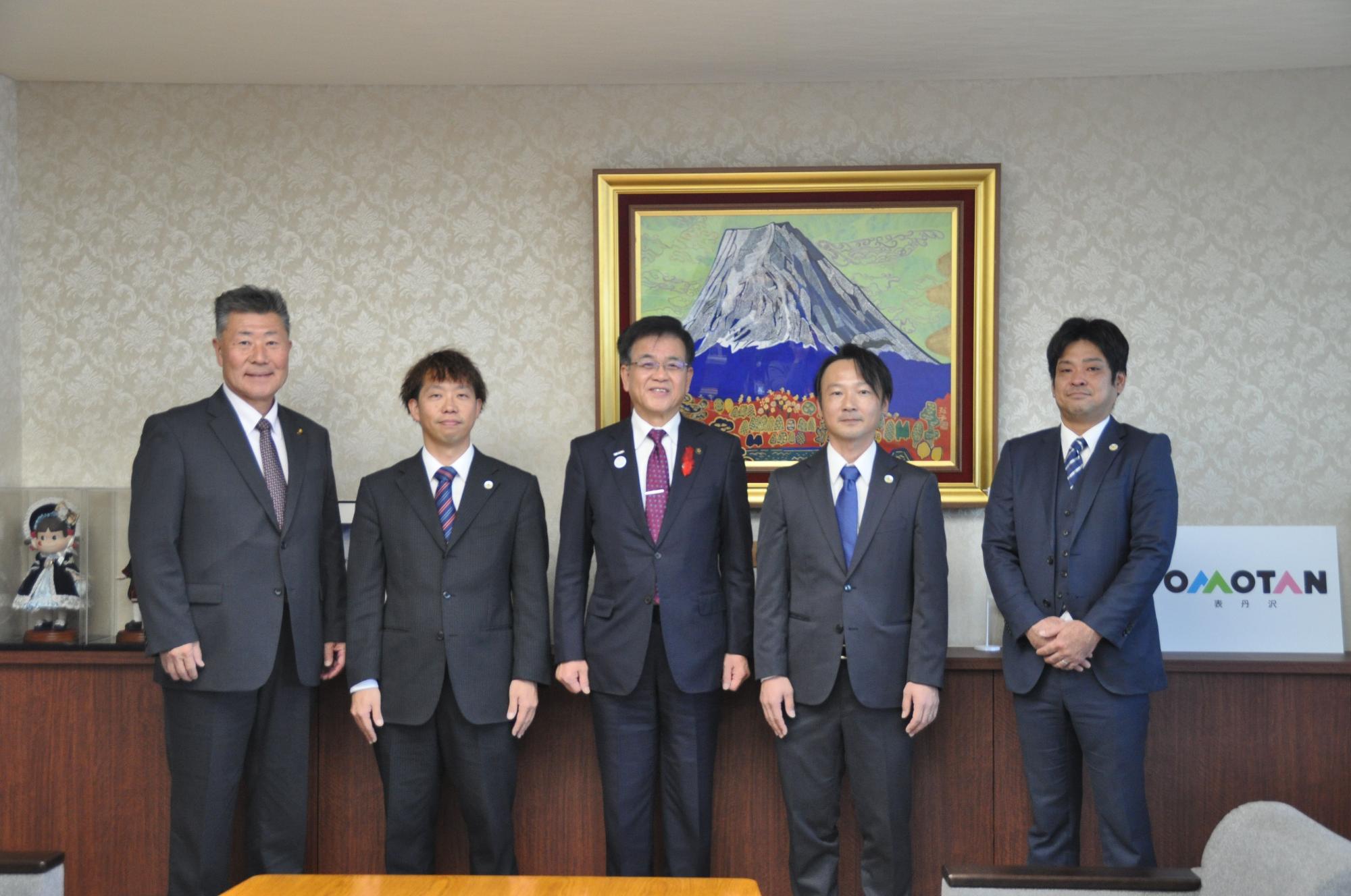 山の絵画が掛けられた壁を背に、高橋市長を中心に、関係者の男性4人が並んで立っている記念写真