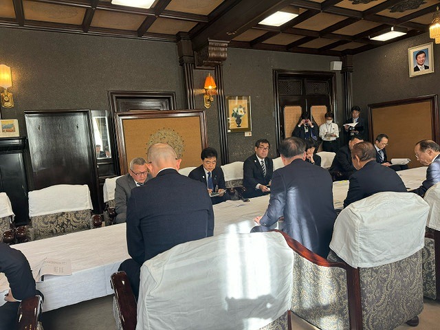 重厚な趣の室内で、白いテーブルクロスが掛かった長いテーブルの両側に配置された椅子に、複数の男性が座り話をしている様子の写真