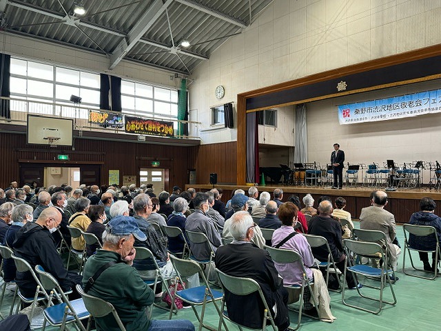 体育館に並んだ椅子に大勢の出席者が座り、舞台の上では市長がスピーチをしている会場全体の様子を後方から撮影した写真