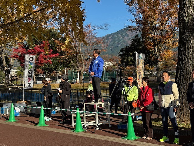 公園で行われたマラソン大会の開会式で、台の上に立ってマイクで挨拶している市長を、複数の人々が囲んでいる屋外写真