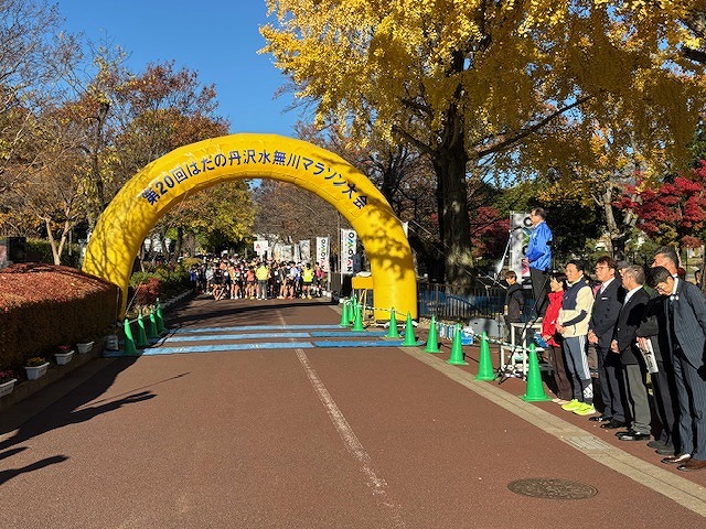 「第20回はだの丹沢水無川マラソン大会」と書かれた黄色いアーチのスタート地点に、たくさんのランナーが並んでおり、横には関係者らしき人々が立っている屋外写真