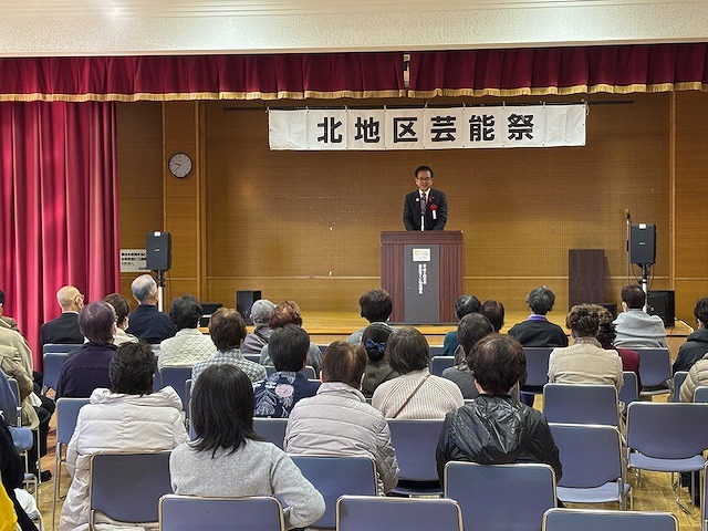「北地区芸能祭」と書かれた横断幕が掲げられた舞台上で、演台に立つスーツ姿の男性が、椅子に座った多くの来場者を前に話しているイベント風景の写真