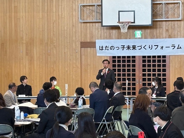 「はだのっ子未来づくりフォーラム」と書かれた横断幕が掲げられた体育館で、マイクを持って話す市長を、多くの参加者が囲んでいるイベント風景の写真