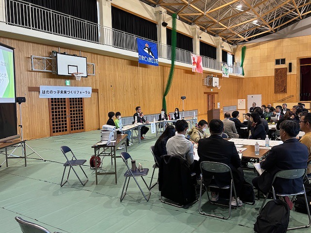 体育館で「はだのっ子未来づくりフォーラム」という横断幕の下、関係者とテーブル席の参加者が議論している様子の写真