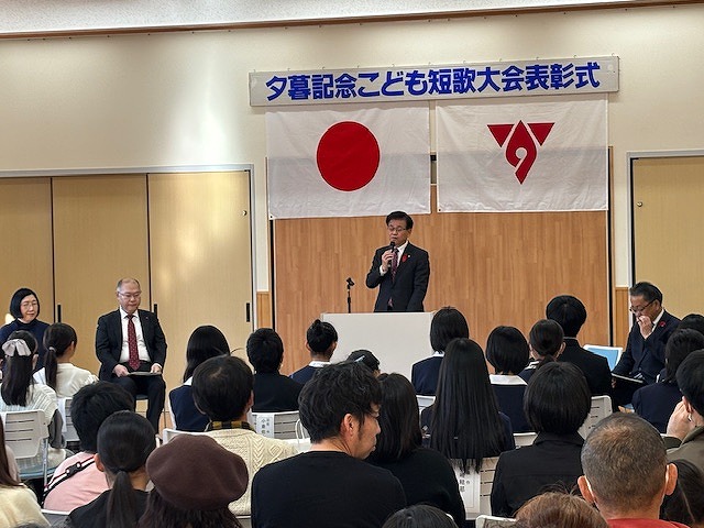 「夕暮記念こども短歌大会表彰式」で、舞台上の演台に立つ市長がマイクを使って挨拶しており、その前に座った多くの来場者が話を聞いているイベント風景の写真