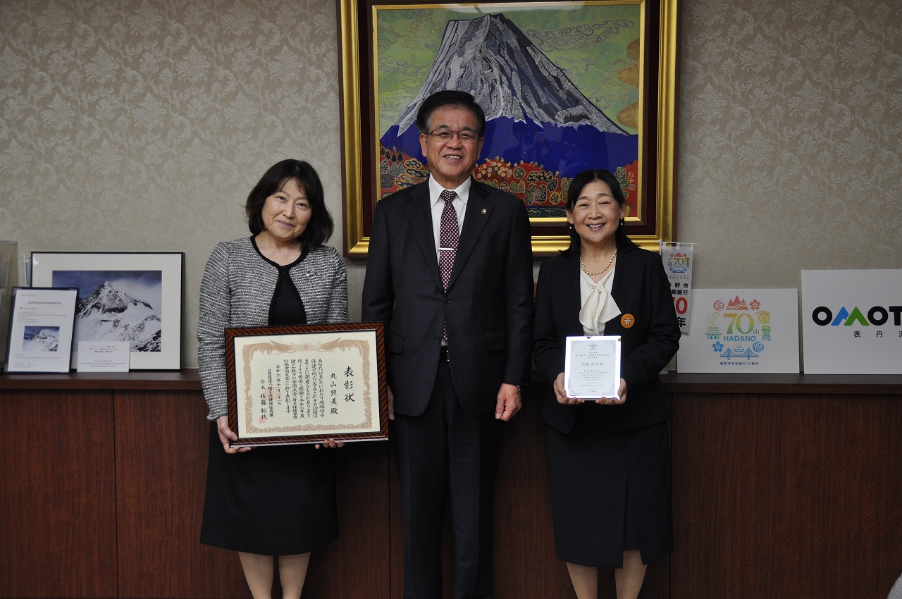 室内で市長と2人の女性が、それぞれ額縁入りの賞状と盾を持って並んでいる記念写真