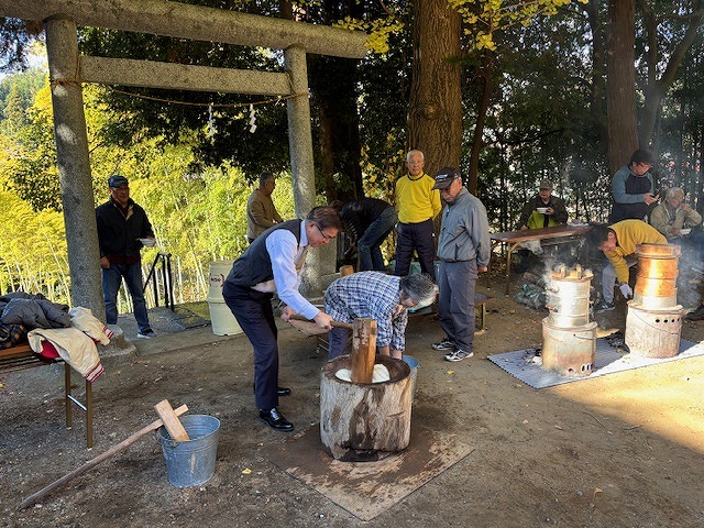 鳥居の下で数人の男性が杵とうすを使って餅つきをしており、周りでは他の人々がそれを見守っている屋外イベントの写真