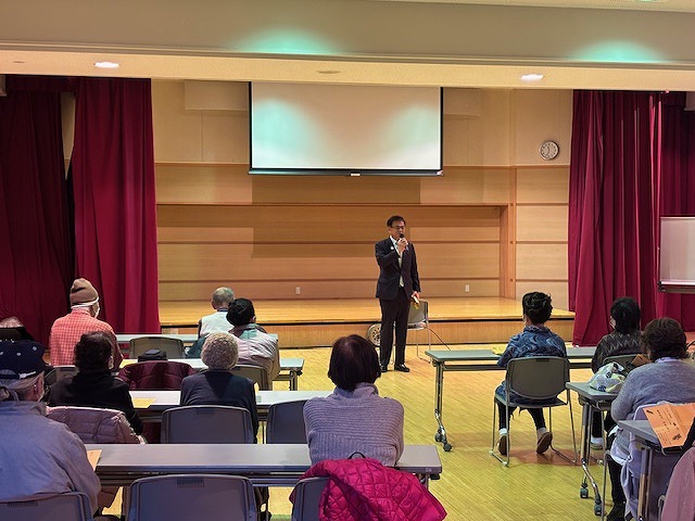 室内講堂のステージ上で、マイクを持って話す市長を、椅子に座った多くの人々が聞いている講演会の写真