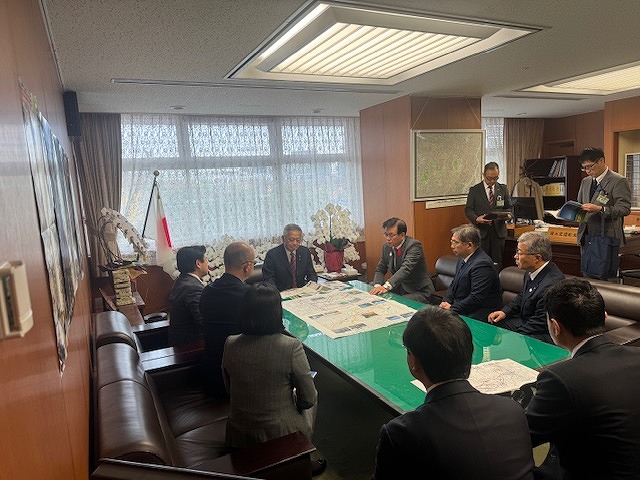 役所の一室と思われる場所で、円卓を囲んで複数の男女が座っており、テーブルに広げられた地図のような資料を見て会議をしている様子の写真