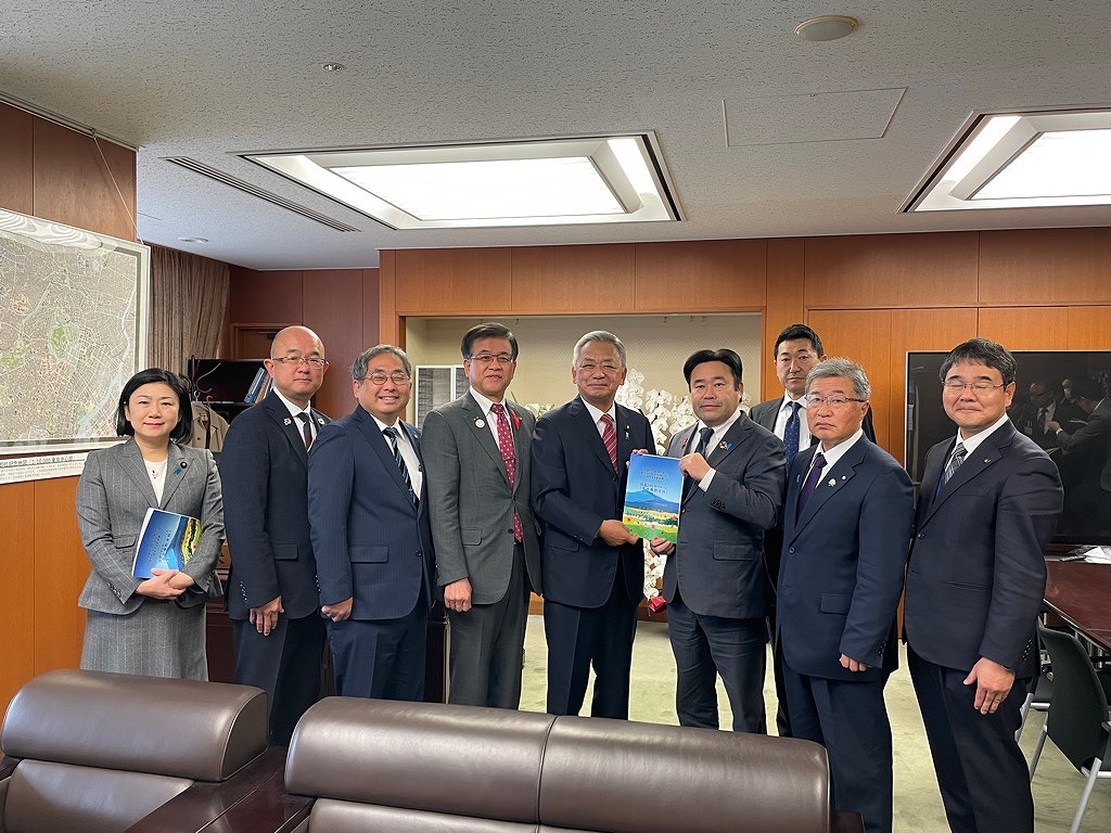 室内でスーツを着た9人の男女が並んでおり、中央の2人が冊子を手に記念撮影をしている集合写真