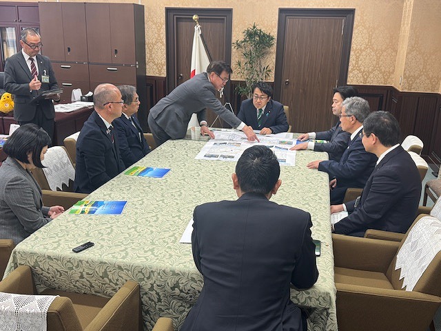 長方形のテーブルを囲む複数の男女が、広げられた資料を見ながら熱心に議論している会議風景の写真