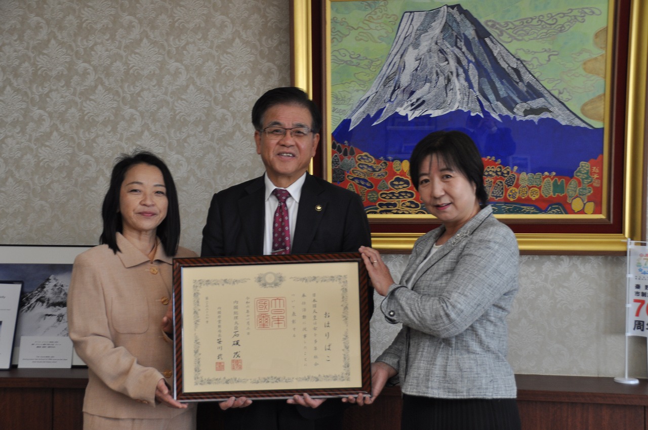 市長を中心に2人の女性が、額縁入りの大きな賞状を一緒に持って記念撮影をしている写真