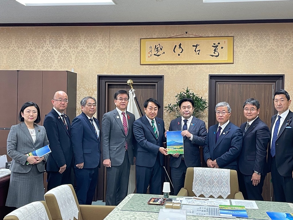 室内で、複数の男女が並んでおり、中央の2人が冊子を一緒に持って記念撮影をしている集合写真