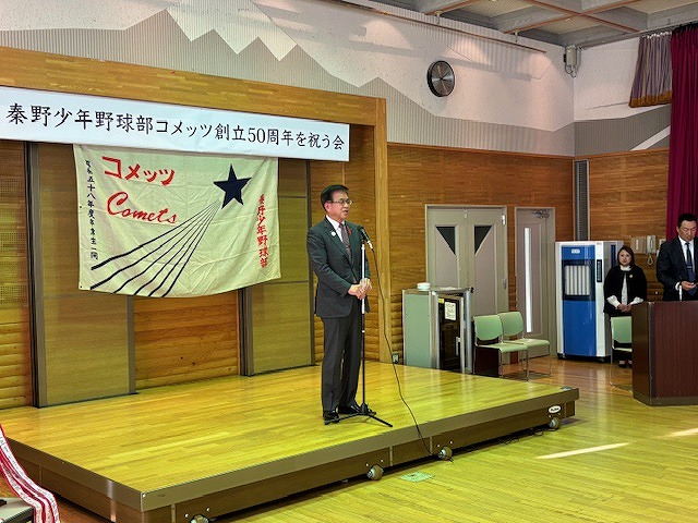 秦野少年野球部コメッツ創立50周年を祝う式典で、舞台上で市長がマイクを使って挨拶しているイベント風景の写真