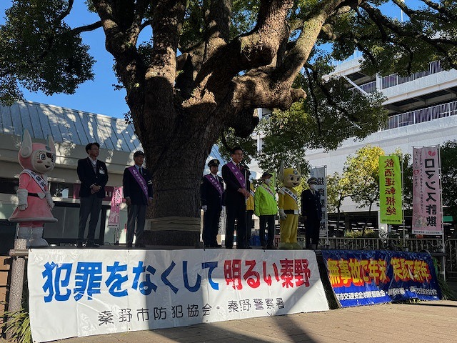 「犯罪をなくして明るい秦野」と書かれた横断幕が掲げられた屋外の舞台で、数人の男性と2体のゆるキャラが並び、そのうちの一人がマイクを持って話しているイベント風景の写真