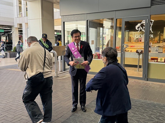駅前の広場で、「秦野市」と書かれた紫色のタスキをかけた市長が、通行人にチラシを配っている屋外活動の写真