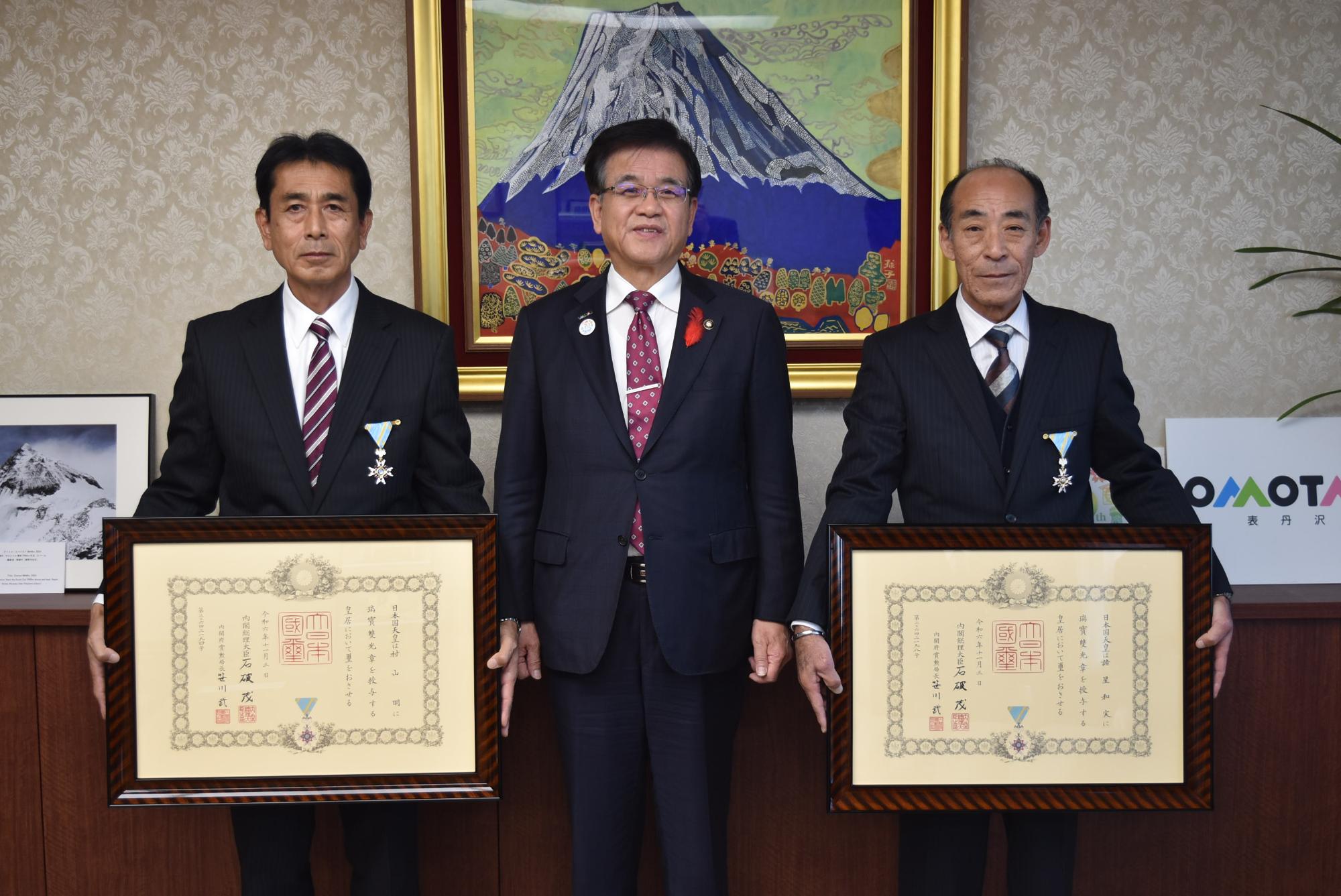 市長と両隣に胸章をつけたスーツ姿の男性が並んでおり、両側の2人が額縁に入った大きな賞状を手に持っている記念写真