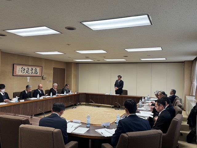 広々とした会議室で、ロの字型に並んだテーブルを囲んで複数の人々が着席しており、中央に立つスーツ姿の男性が挨拶をしている一市三町広域行政推進協議会の写真