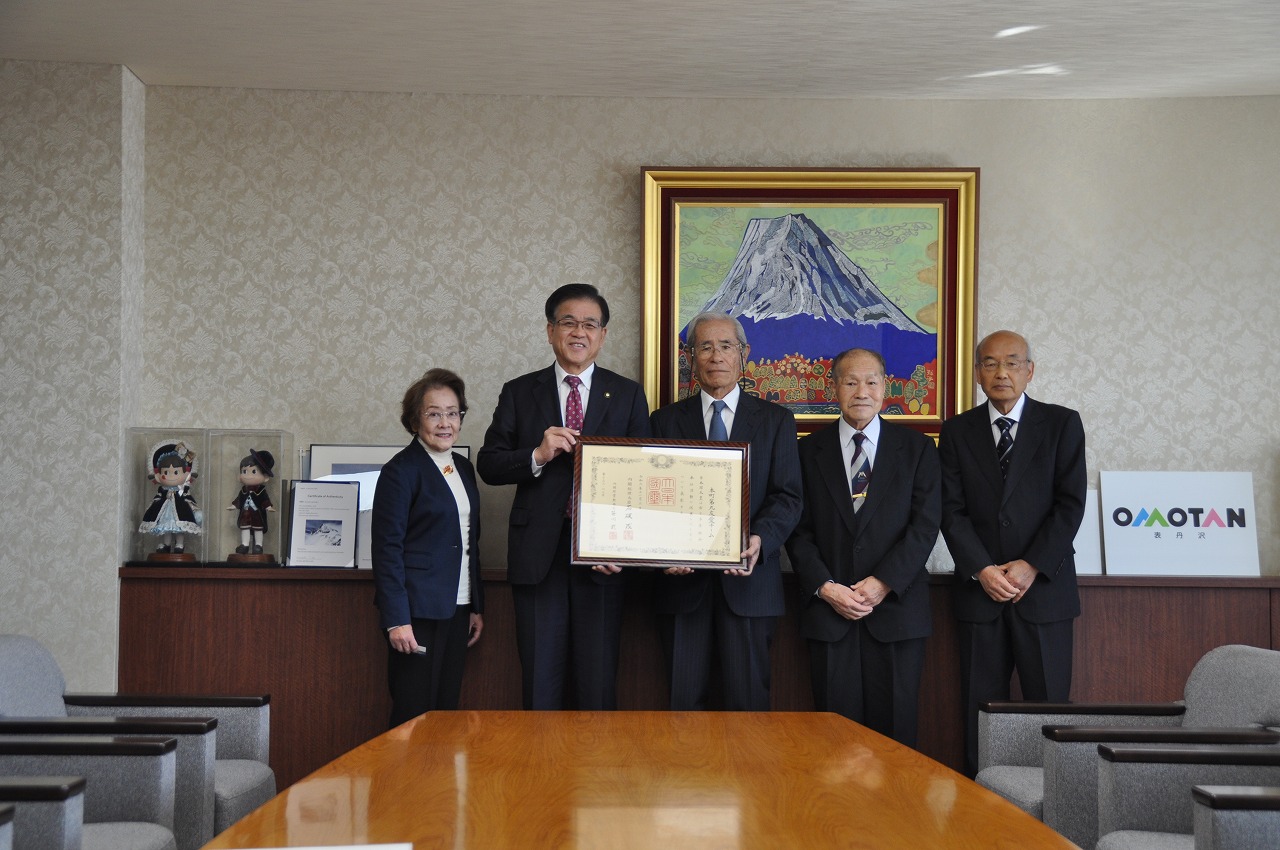 室内で市長を含めた5人の男女が並んでおり、そのうちの2人が額縁に入った大きな賞状を一緒に持って記念撮影をしている写真