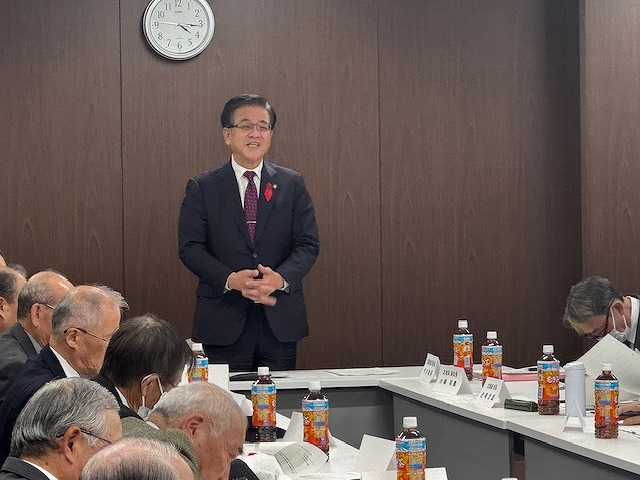 長机を囲んで座る多くの人々を前に、市長が立って挨拶をしている懇談会風景の写真