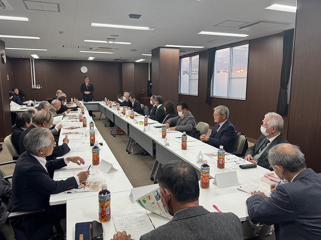 ロ字型に配置されたテーブルを囲んで複数の人々が着席しており、その後方でスーツ姿の男性が立って話している懇談会風景の写真