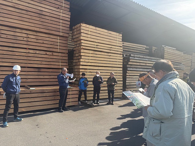 木材が山積みにされた製材所で、メガホンを持った男性が説明をしており、他の参加者が話を聞きながら資料を見ている屋外写真