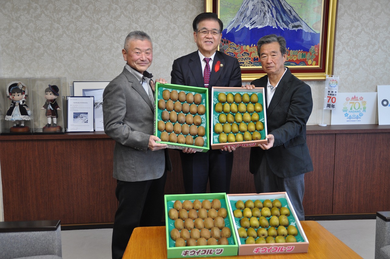 市長を含めた3人の男性がキウイフルーツがぎっしり詰まった箱をそれぞれ手に持ち、カメラの方を向いて立っている記念写真