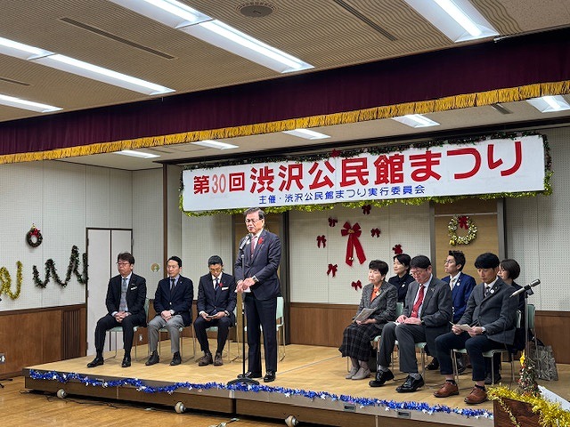 「第30回渋沢公民館まつり」と書かれた横断幕が掲げられた舞台で、マイクを持って話すスーツ姿の市長を複数の関係者たちが囲んでいるイベント風景の写真