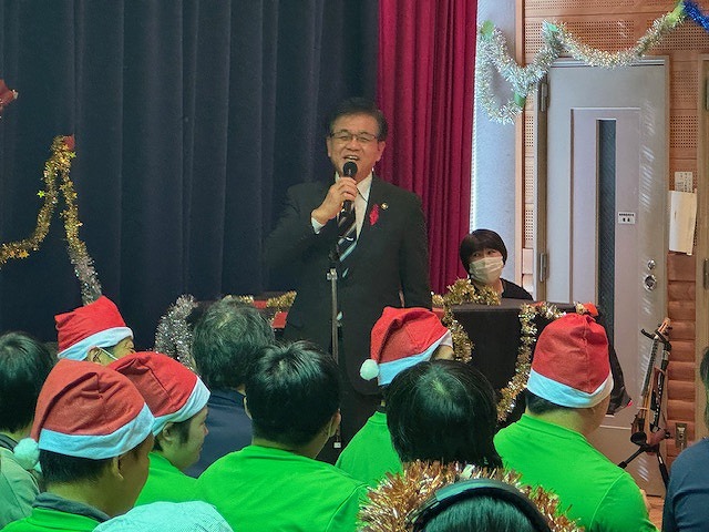 赤いサンタ帽子をかぶった大勢の人々を前に、市長がマイクを持って挨拶している、クリスマスイベントの様子の写真