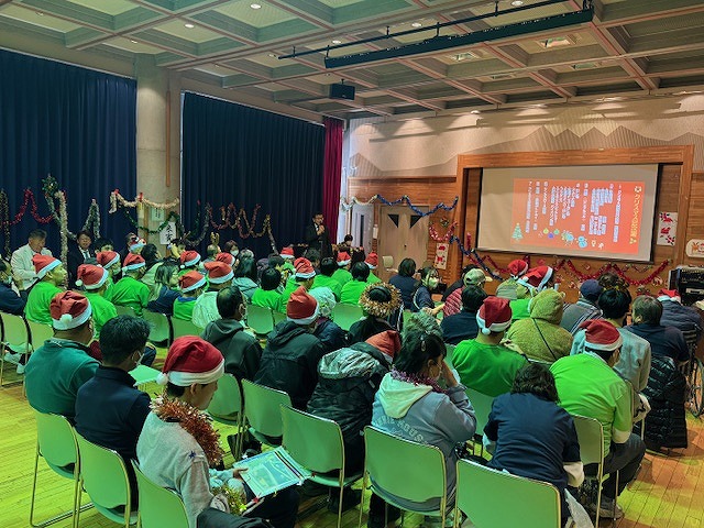 体育館で行われたクリスマスイベントで、サンタの帽子をかぶった大勢の観客が、舞台上のスクリーンとマイクで挨拶する男性に注目している様子の写真