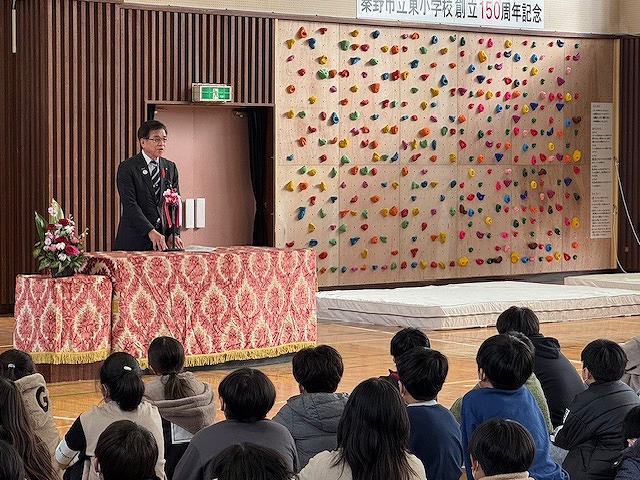 背景にボルダリングウォールが見える体育館内での記念式典で、演台に立つスーツ姿の市長が、たくさんの生徒が座っている前で挨拶をしている写真